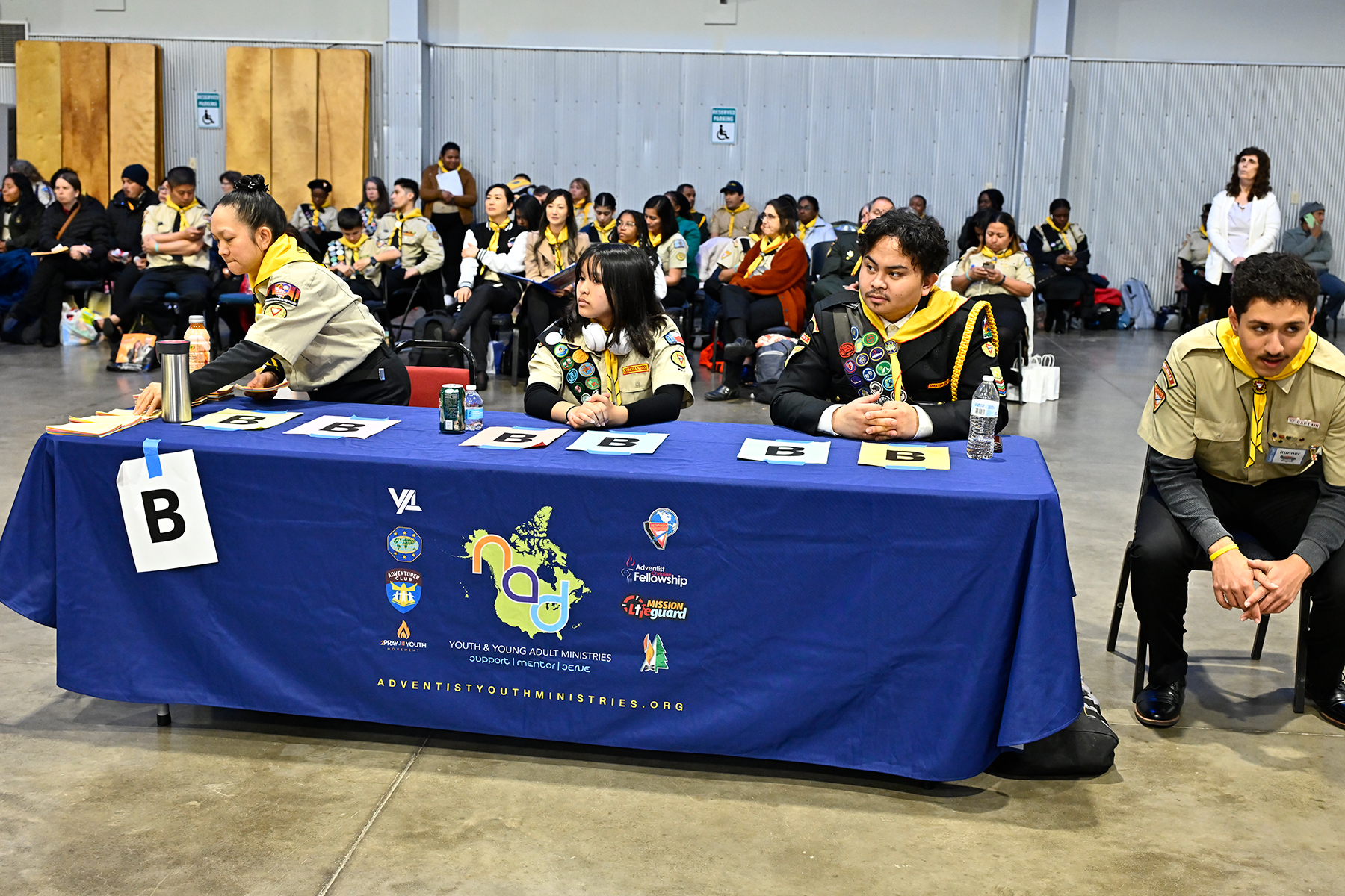 People in Pathfinder uniforms sort papers, while a large crowd of people sit behind them.