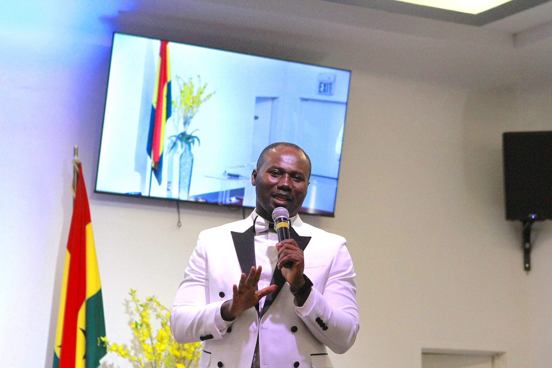 Black man preaching with a mic in hand. The Ghanaian flag is to the left of him and a screen showing another angle of the flag behind him.