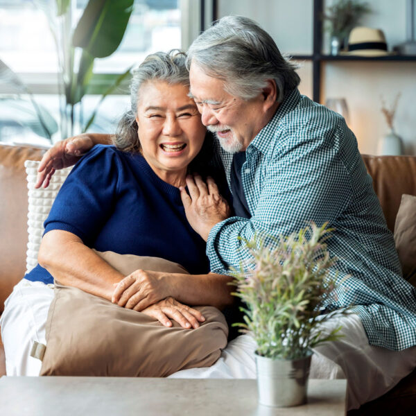 Older Asian couple shares a smile and hug while sitting on a couch.
