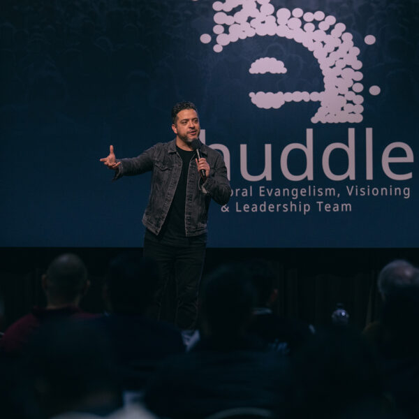 Hispanic man speaking on a stage with a screen reading eHuddle behind him.