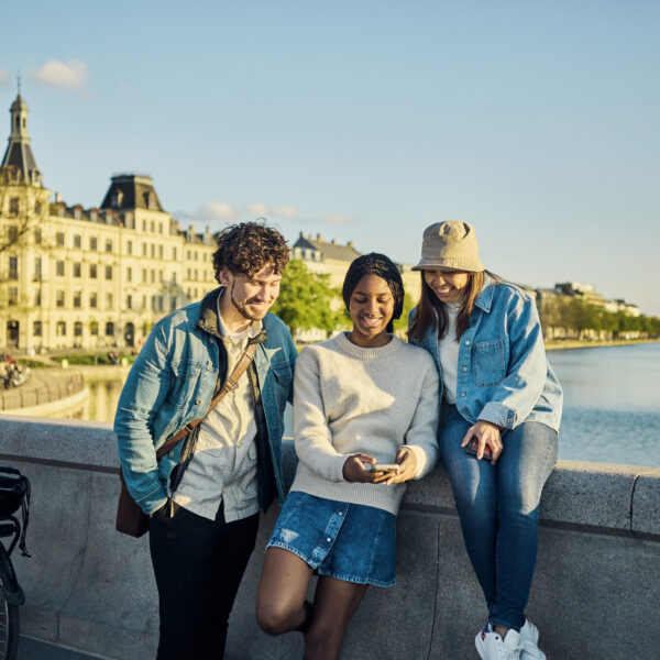 Three college age friends in Europe sit on the edge of a bridge in front of a ornate older building along a river.