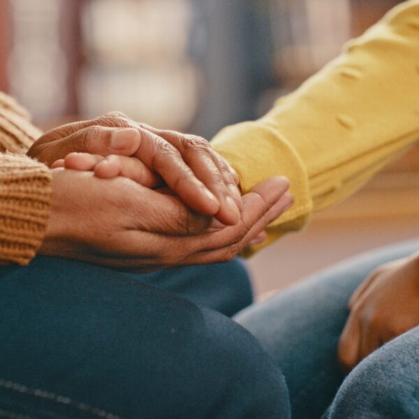Close up photo of two people sitting knee to knee with one clasping the hand of the other to comfort them.