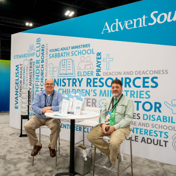 Two men sit on stools by their business display at a convention exhibit hall.