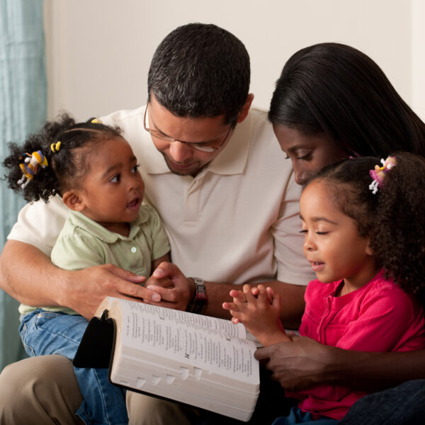 Young family with two children praying together at home on their couch. Toddler has eyes open and is looking at her mother.