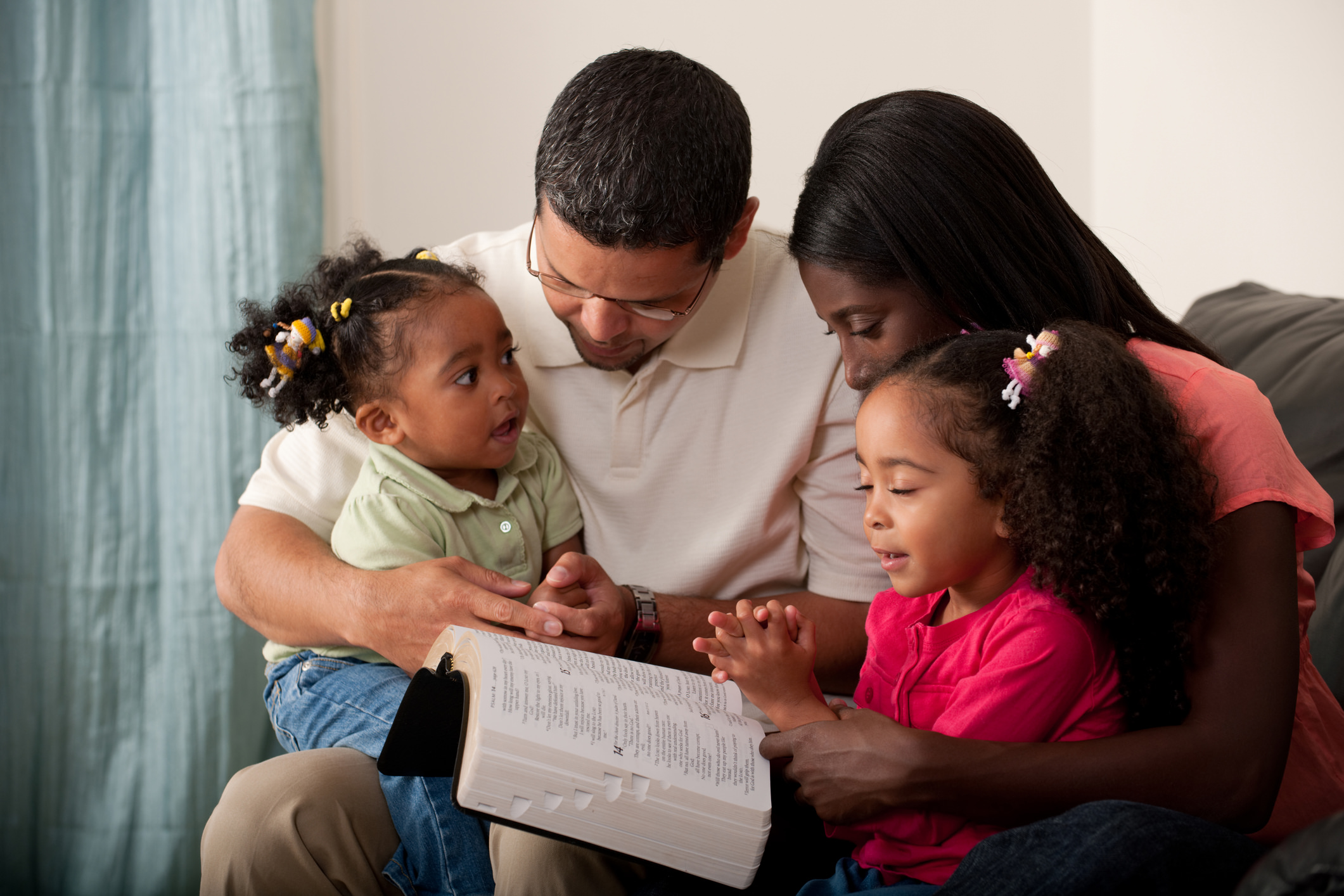 Young family with two children praying together at home on their couch. Toddler has eyes open and is looking at her mother.