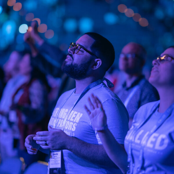 Photo focuses on two people in a large crowd at an event. People are in blue light from the stage and have their eyes closed and hands raised.