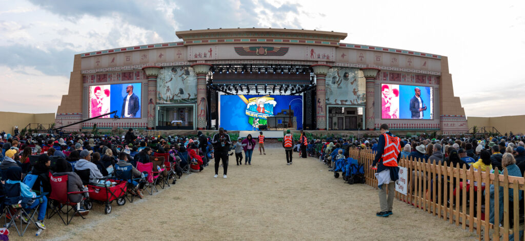 Photo showing a large outdoor stage for a religious yout program at dusk and many people sitting on camping chairs making up the audience.