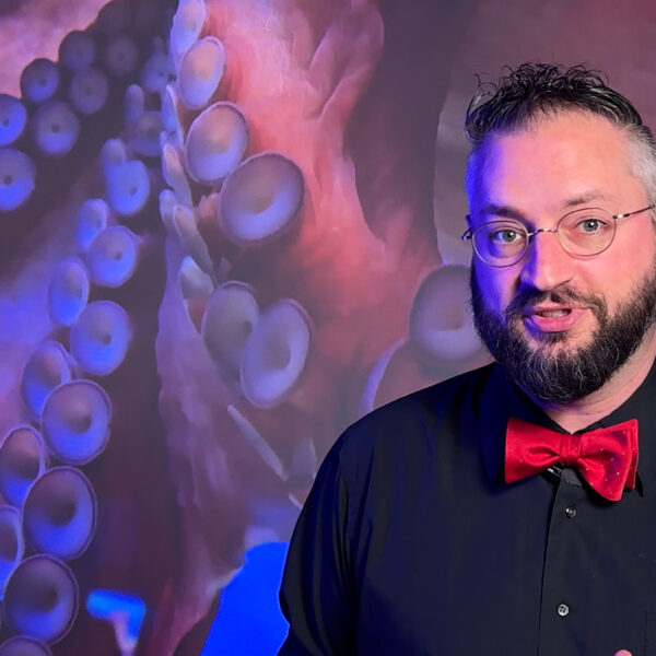 Caucasian man in black dress shirt and red bowtie poses in front of octopus picture