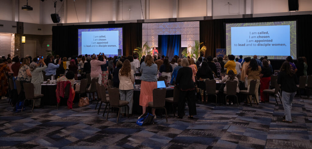 A large group of women stand in a conference hall during a session. Two screens at the front display the words, ‘I am called, I am chosen, I am appointed to lead and to disciple women.'