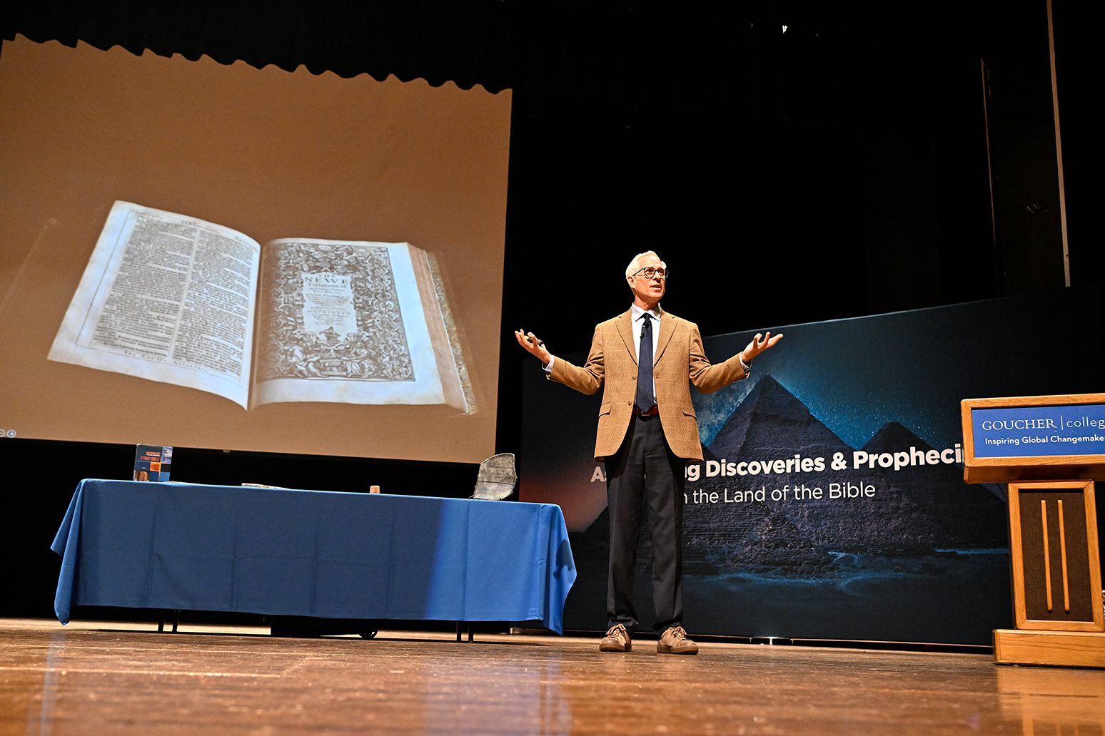 A man stands on stage beside a large slide of a Bible.