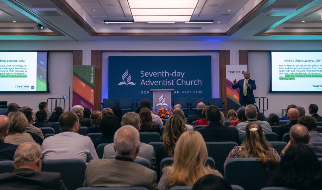 Photo of a large business meeting with a seated crowd. Taken from the back and towards the front with a presenter and screens.