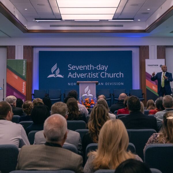 Photo of a large business meeting with a seated crowd. Taken from the back and towards the front with a presenter and screens.