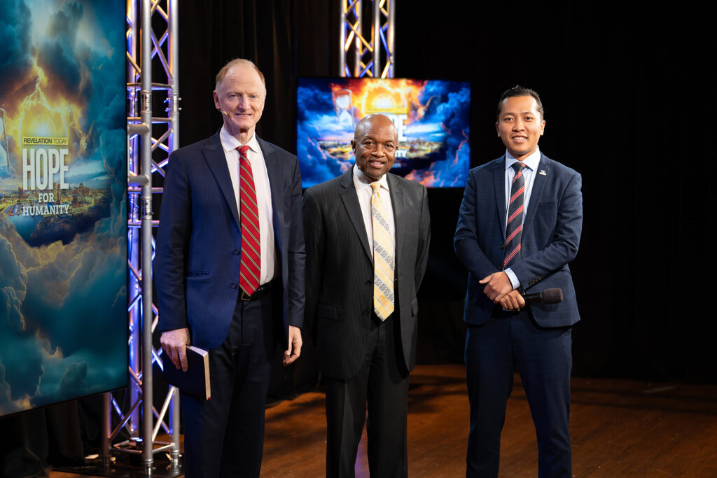 Three men stand on a stage in front of a banner and TV screen emblazoned with the Hope for Humanity logo