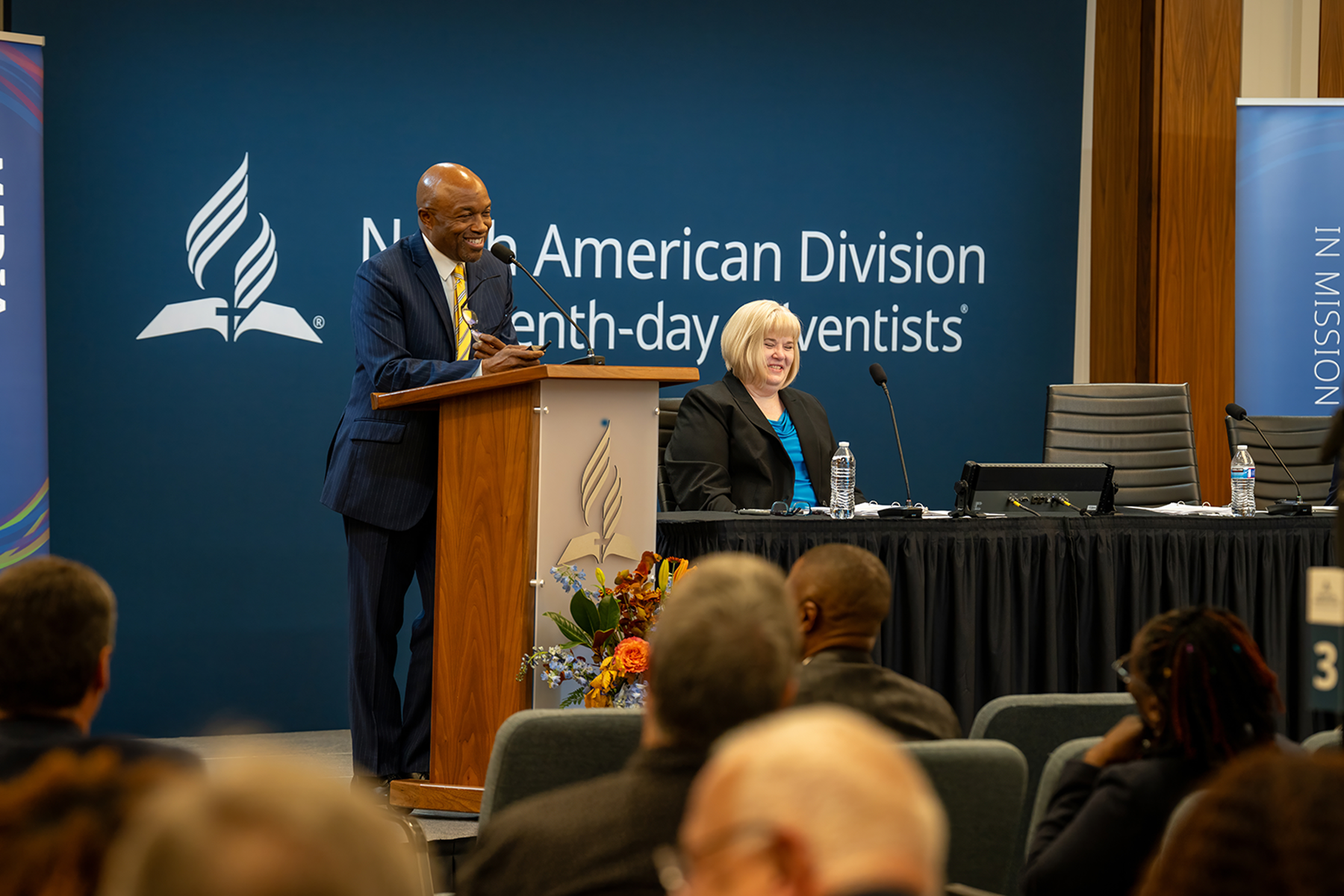 A man speaks at a podium during a meeting, smiling. A woman sits beside him at the front table, with the NAD logo displayed on the wall behind them.