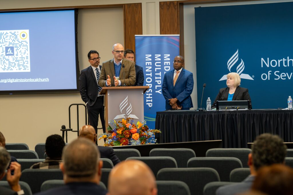 A man speaks from a podium while three other men stand nearby and a woman sits at a table beside him.