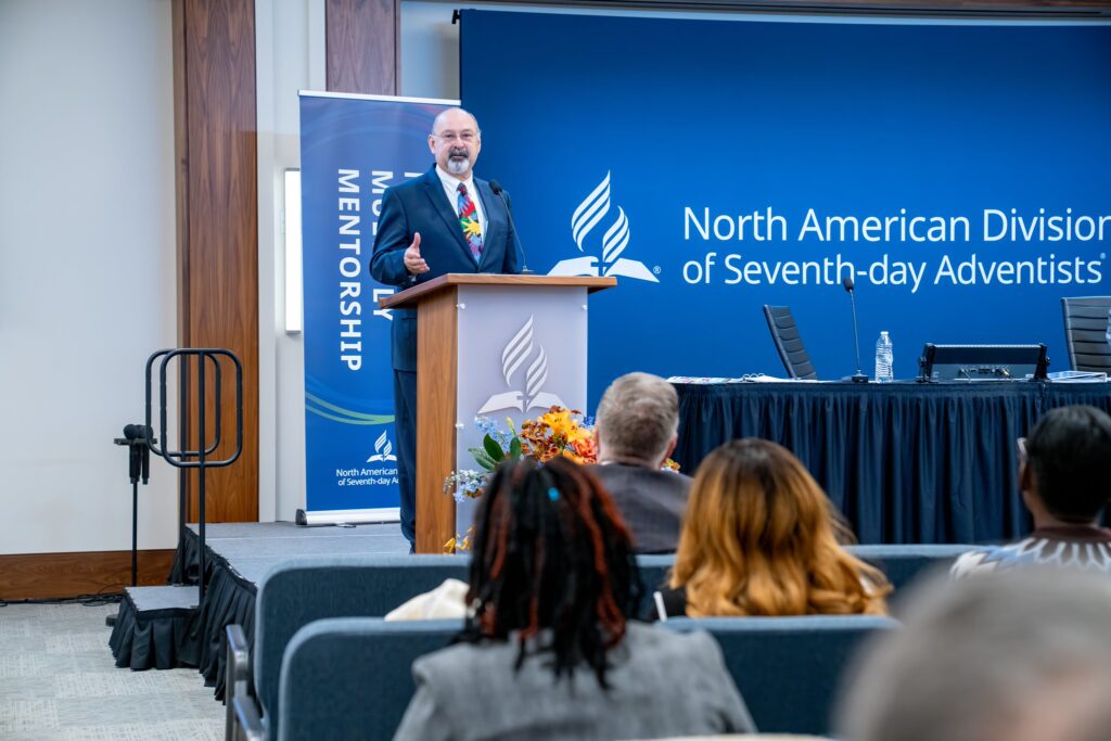 Older white man in a suit addressing attendees at a meeting.