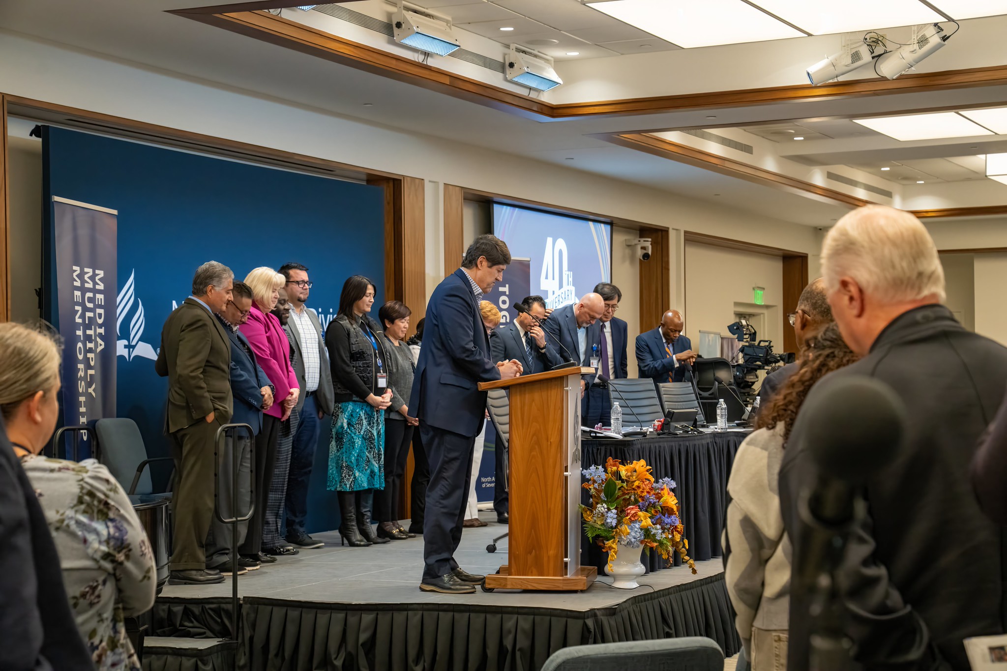Mixed group of business people stand on a stage with heads bowed in prayer.