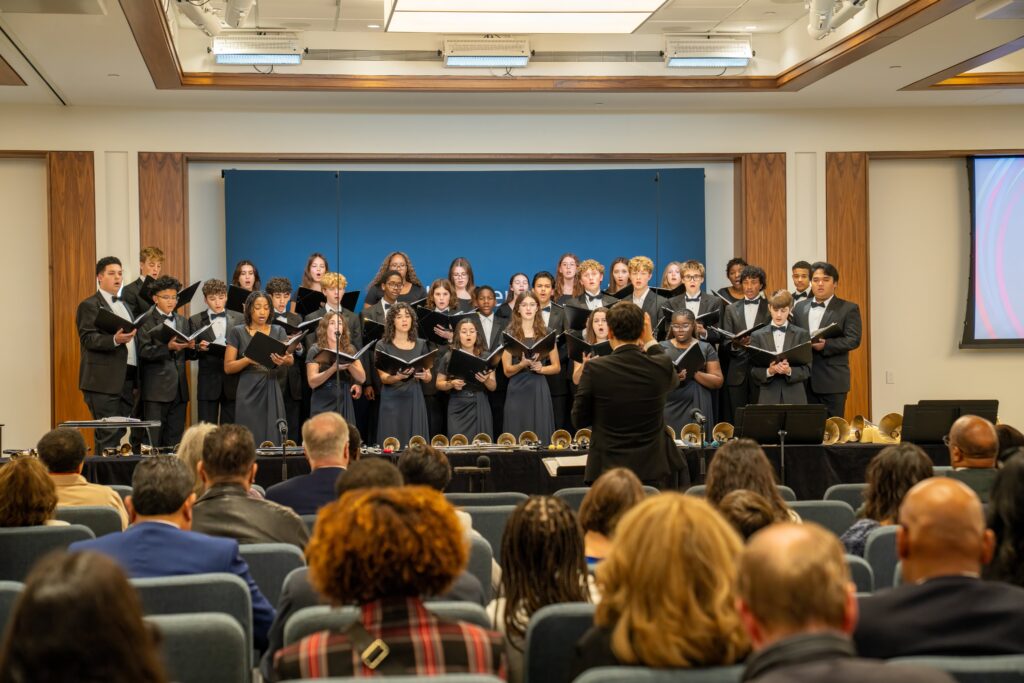 A diverse choir of young people in black sing in front of an audience.