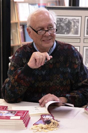 Older caucasian man with glasses signs a copy of his book at a book signing