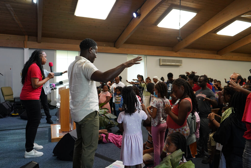 A woman in a red t-shirt prays in a sanctuary filled with several children and adults. 