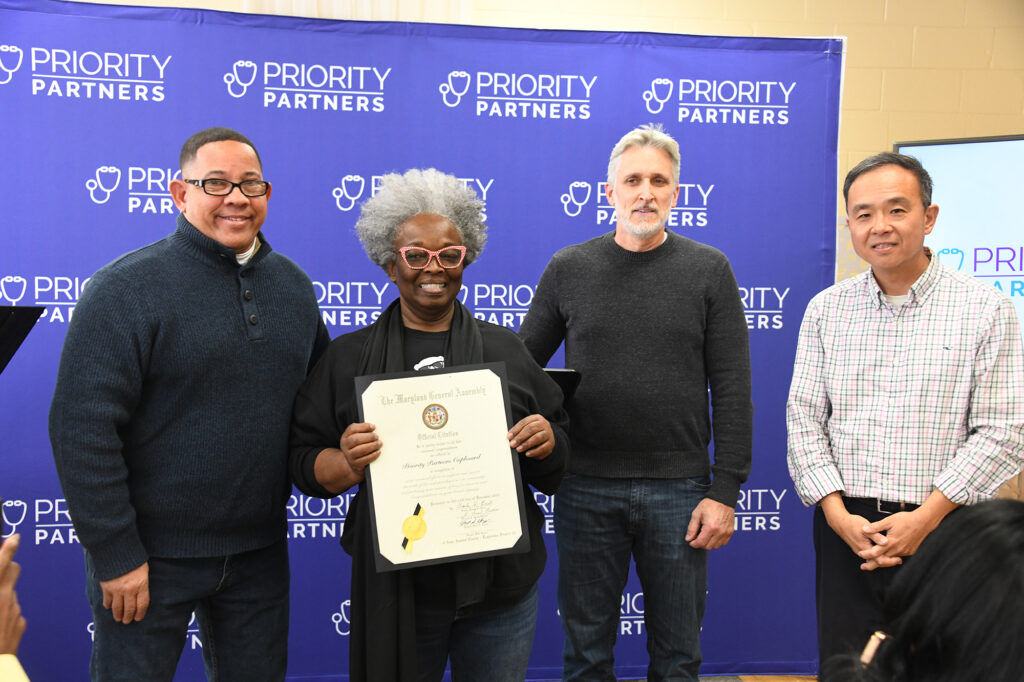 Three men and a woman stand in front of a purple backdrop, the woman holding a certificate. 