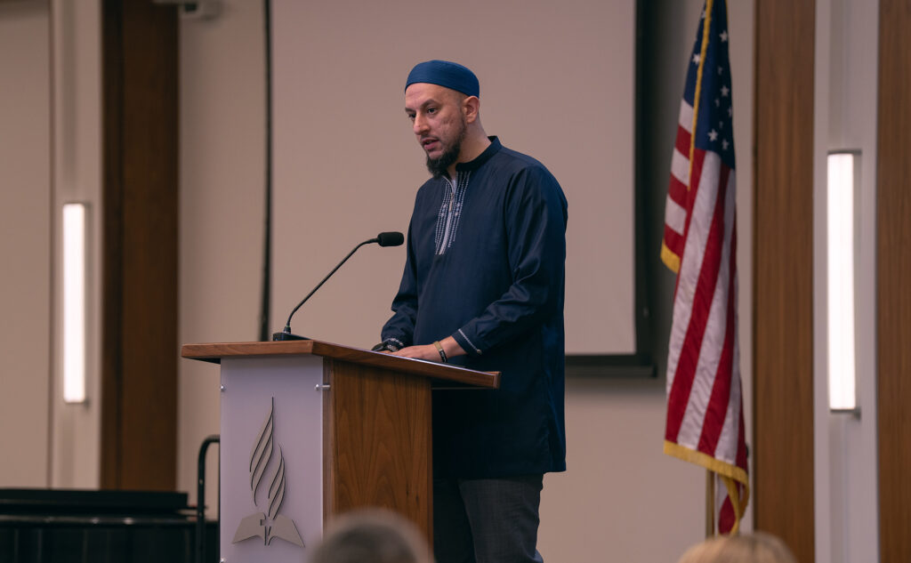 A man in an Islamic tunic stands at the podium, the American flag in the background.