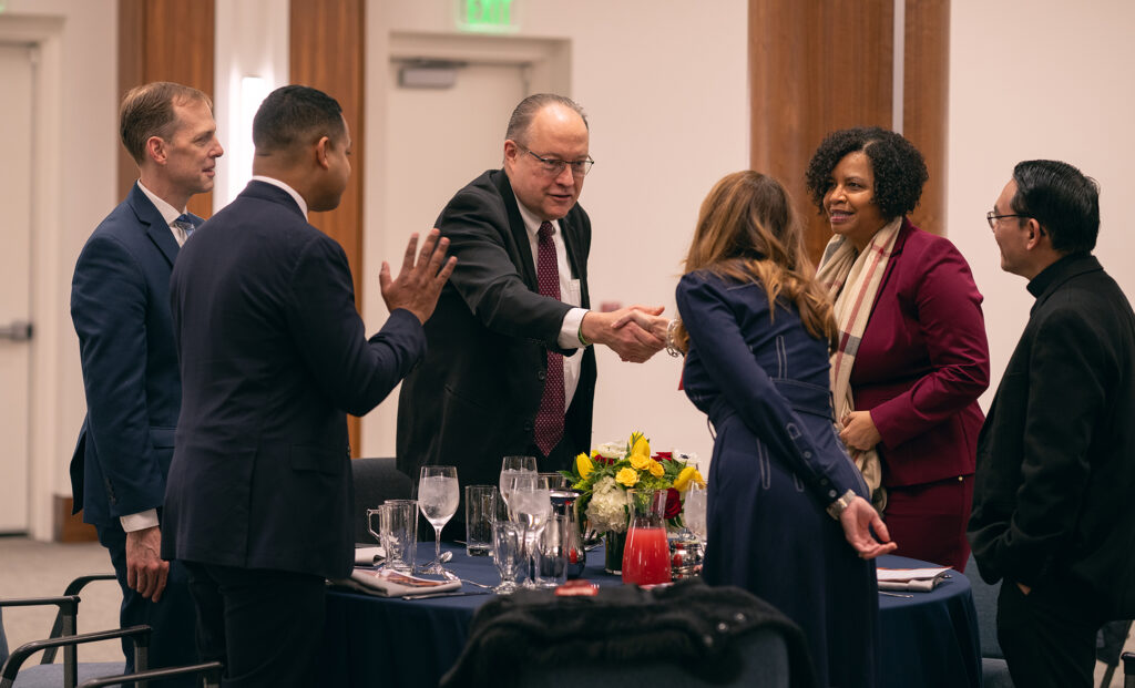 A dvierse group of men and women stand around a round dining table meeting and greeting.