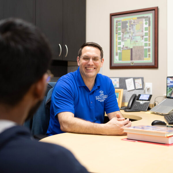 A male college professor speaking with a male college student in his office.