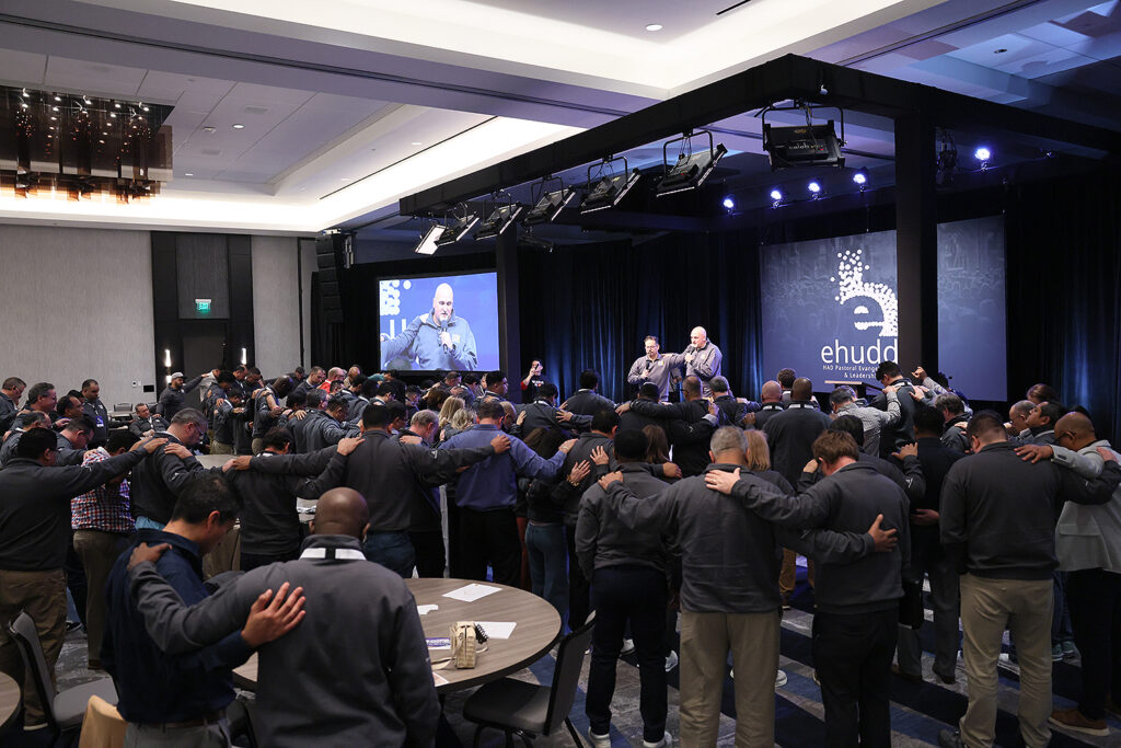A large group of pastors huddle together near a stage.