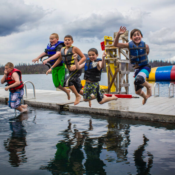 Several boys jump off a deck at a lake.