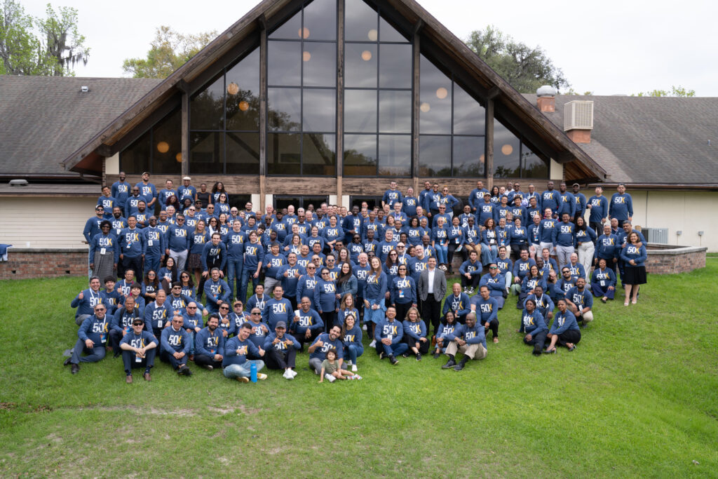 A group of participants in blue shirts gathers behind a building for a group photo.