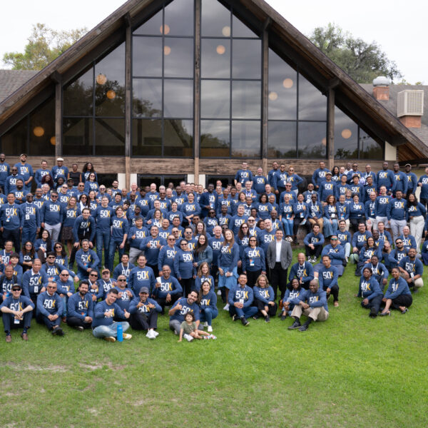 A group of participants in blue shirts gathers behind a building for a group photo.