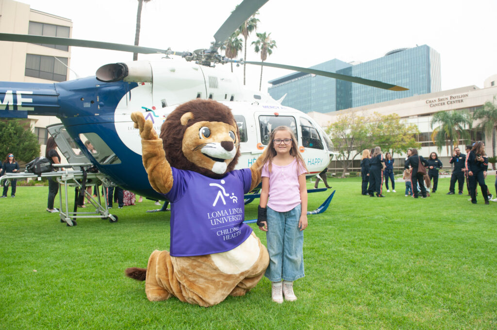 A young girl stands with a teddy bear mascot stands in front of Loma Linda University's new pediatric transport helicopter.