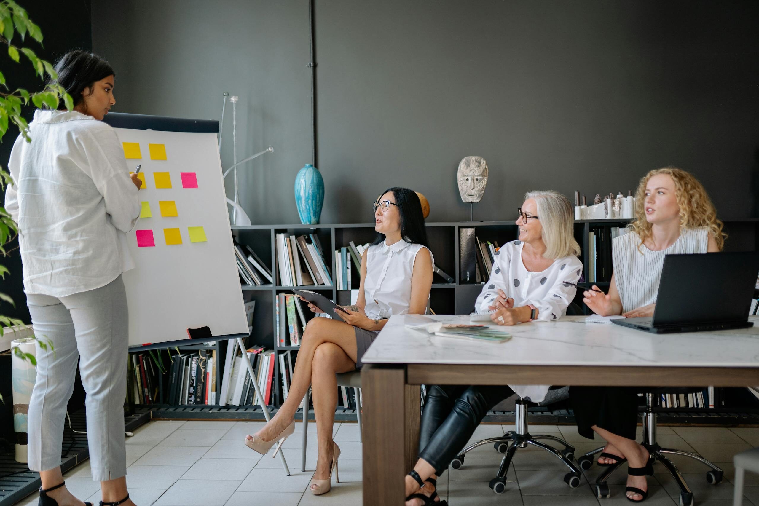 A diverse group of women collaborating in an office setting, discussing ideas using a flip chart and laptop.