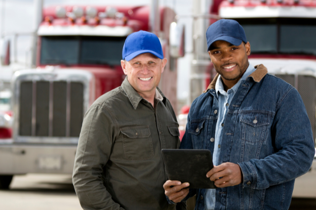 Two truck drivers - one holding a clipboard - are standing in front of a red commercial truck and smiling at the camera.