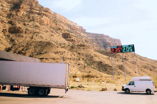 A tractor-trailer and a camper van are parked under a gas station price sign in front of a desert cliff.