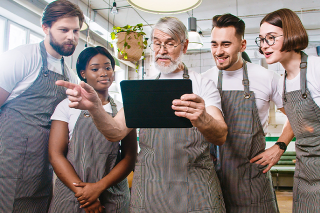 Four restaurant employees and their manager gather in the kitchen to discuss plans for the upcoming shift. Each person is wearing an apron and looking at the screen of a shared tablet. 