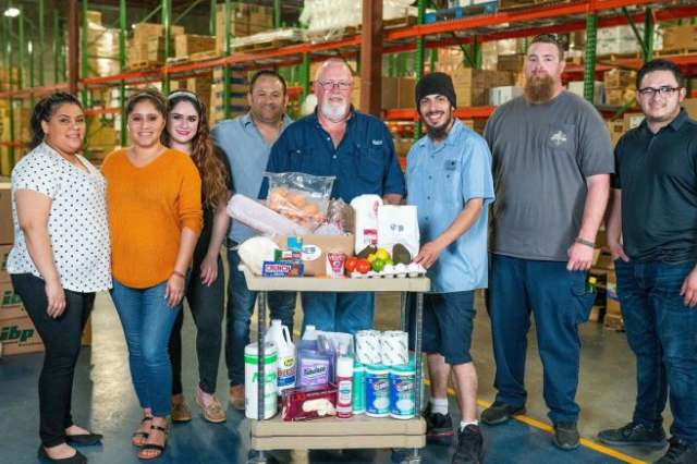 A group of employees are gathered around a collection of food service products in a warehouse. 