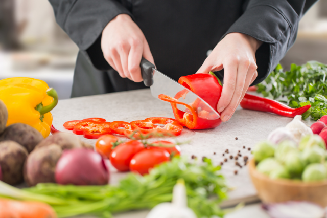 Close up of a chef slicing a red bell pepper on a counter covered with various ingredients.