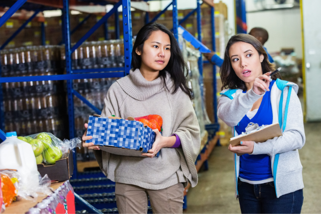 A woman holding a clipboard pointing another woman carrying a box full of fruit to something in a warehouse