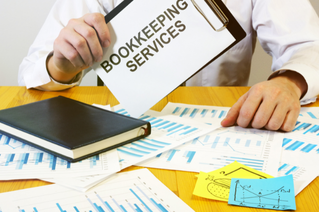 A person sitting at a desk with papers and books. They’re holding a clipboard that says “bookkeeping services.”