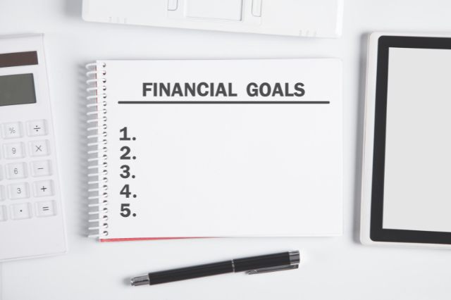 A notebook on a desk surrounded by a pen, calculator, and other objects. The notebook says Financial Goals with a list with the numbers 1-5 written out. 